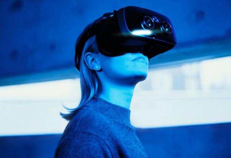 Kuvassa on nuori nainen mustat virtuaalilasit päässään puolivartalokuvassa. Taustalla on pitkä kapea ikkuna.  Kuvan sävy on sininen. 