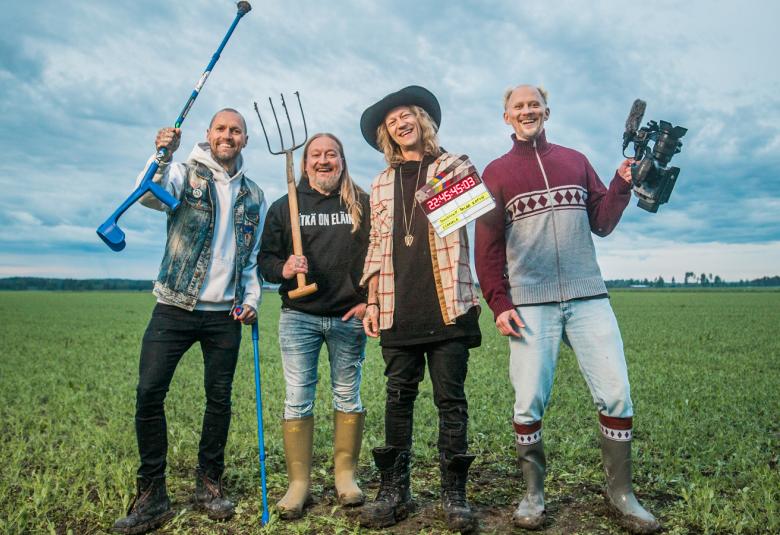 Kuvassa on nurmipellolla neljä Duudsonin jäsentä college-paidoissa ja Jussi-paidassa kädessään hanko tai muita työvälineitä. Taustalla on sinertävää pilvitaivasta.