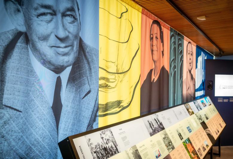 Kuvassa on vasemmalla rivi kuvia näyttelystä seinällä ja kuvat ovat sinisiä, keltaisia, punaisia ja vihreitä. Alla lasivitriinit.