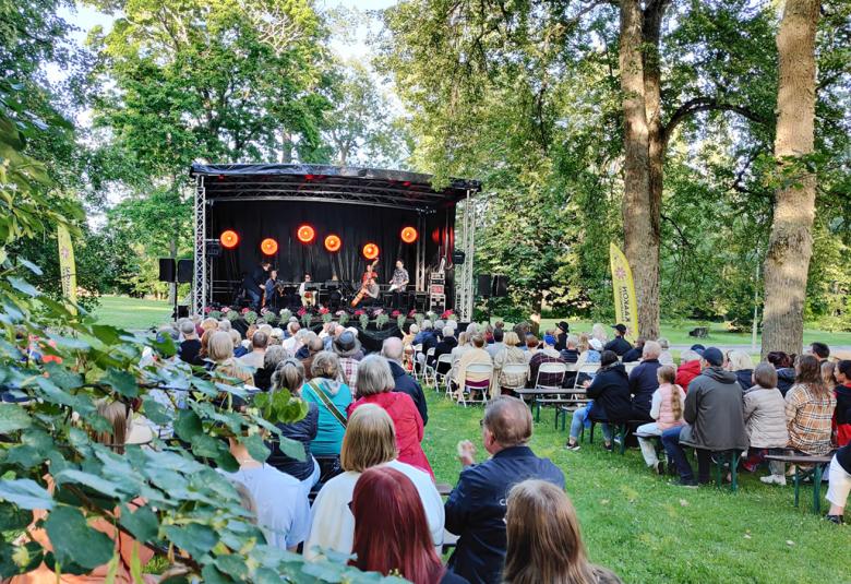 Kuvassa on takana konserttikatos, jossa on viulisteja ja kontrabasson soittaja. Vasemmalla ja oikealla on yleisöä puistossa vihreällä nurmikolla ja lehmuksen alla selin.