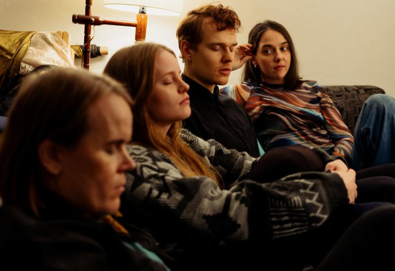 Kuvassa ovat Camille Auer, Anna Rosaliina Kauno, Samuel Kujala, Kaisaleena Koskenkorva istumassa vinottain sohvalla ja kuva on puolivartalokuva.  He näkyvät melko tummina. Taustalla on kellertävää valoa.