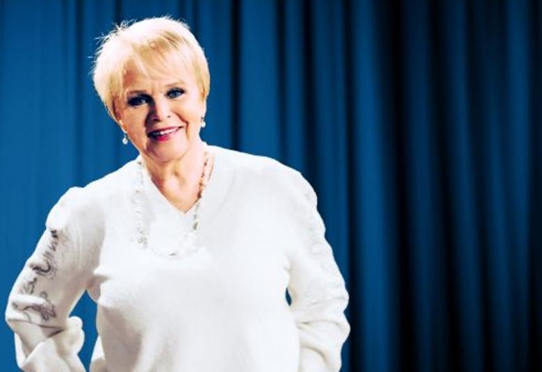 Kuvassa on Katri Helena puolivartalokuvassa seisomassa ja hänellä on valkoinen villäpaita päällään sekä helmet. Taustalla on kirkkaan sininen verho.