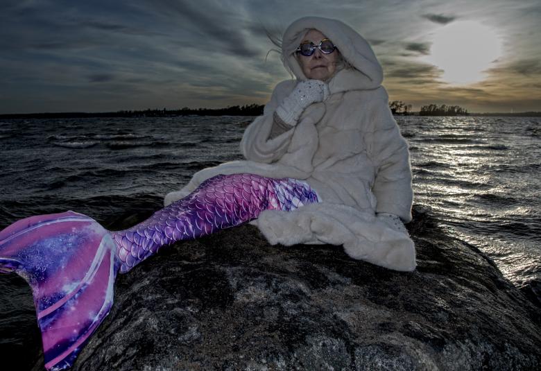 Kuvassa on Seela Sella merenneitona pitkässä pyrstössä kallion päällä ja tausta lla on tummaa merta. Oikealla on täysikuu ja kuun kajo vedessä.