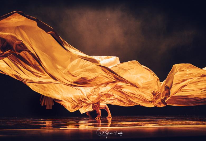 Kuvassa on mustalla taustalla leveästi leijuvaa oranssia kangasta.  Hiukan vasemmalla näkyy tanssijan jalat.