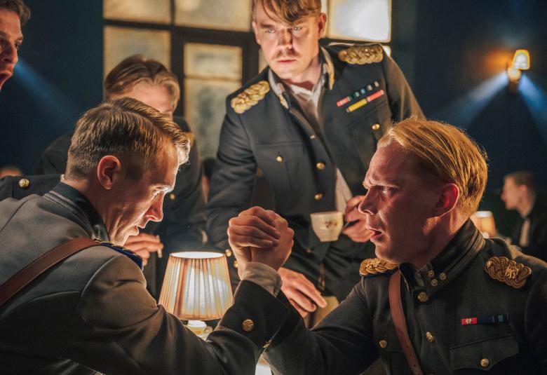 Kuvassa on miehiä tumman vihreissä upseeripuvuissa. Takana seisoo Tom Rejströn ja pöydän ääressä edessä vääntävät kättä Jussi Vatanen ja Pääry Oja.