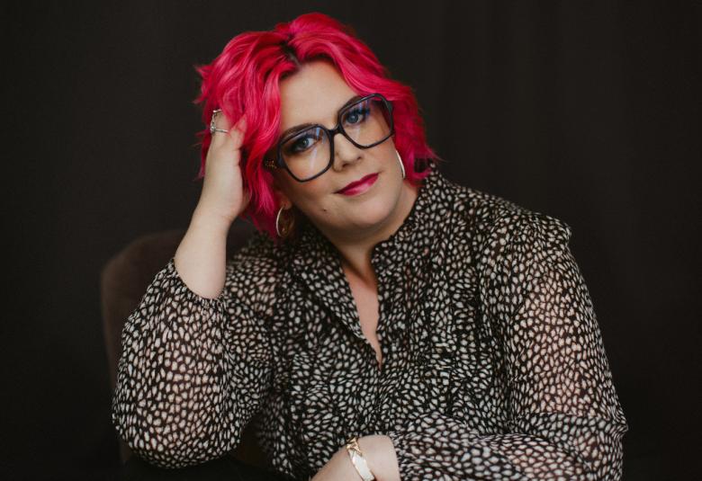 Kuvassa on Nilla Kjellsdotter puolivartalokuvassa ja hänellä on harmaa jakku päällään ja kirkkaan punaiset hiukset. Hän pitää oikeaa kättään hiuksissaan. Tausta on tumma.