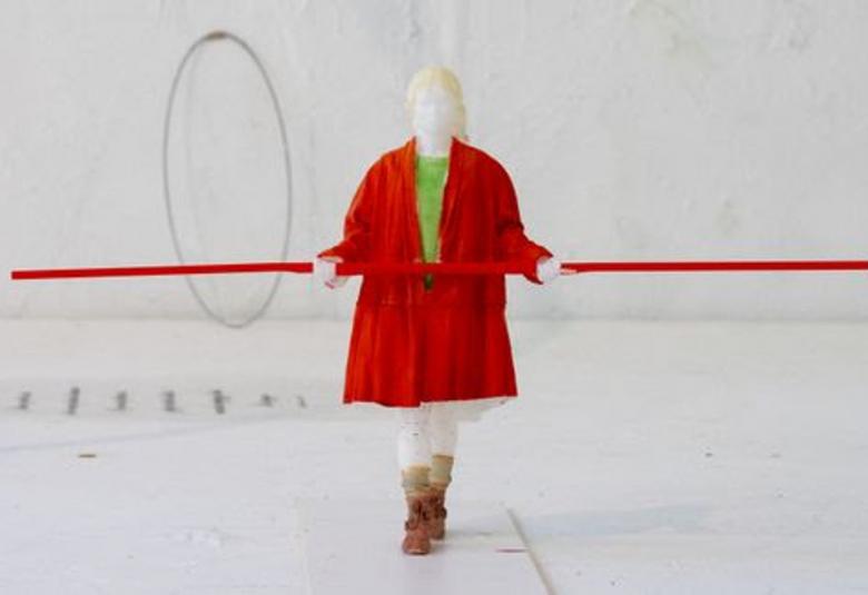 Kuvassa on punaisessa takissa oleva hahmo, jolla on punainen tanko kädessään vaakatasossa. Tausta on vaalea.