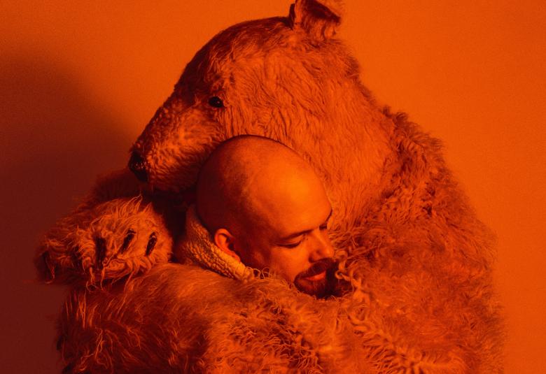  Kuvassa on oranssina koko kuva ja siinä on tanssija Taneli Törmä syleilemässä karhua.