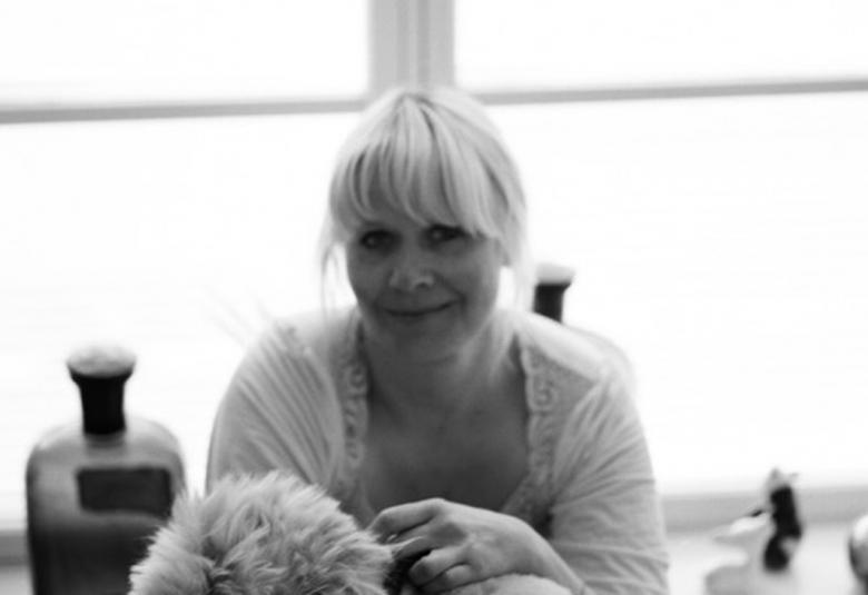 Kuvassa on Paola Suhonen istumassa pöydän ääressä. Pöydällä on lasipulloja ja ikkunalla omena.