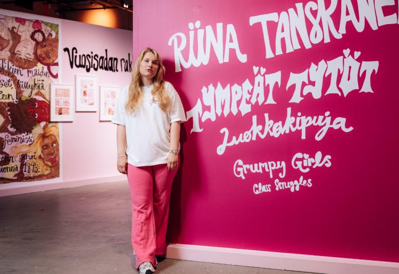 Kuvassa on riina Tanskanen vaaleassa puserossa ja punkeissä housuissa. Veressä on pinkillä seinällä on näyttelyn nimi isoin valkoisin kirjainmin Riina Tanskanen, Tympeät tytöt, Luokkakipuja, Grumpy Girls. Vasemmmalla oikealla takana on pinkin ruskeata teosta. 