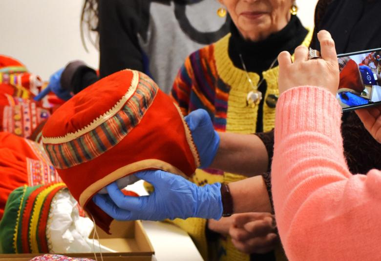 Kuvassa on punaisia saamelaispäähineitä. Oikealla on nainen vaalean punaisessa villatakissa, joka ottaa kännykällä kuvaa. Hänestä näkyy osa vartaloa. Taustalla näkyy nainen siten, että kasvoja ei näy kokonaan. 