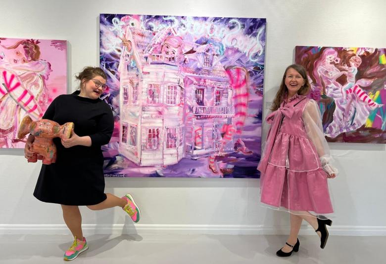 Kuvassa on vasemmalla tauluna pinkkisävyinen tyttö istumassa ja keskellä on violettisävyinen isompi taulu, jossa on vaalea talo. Oikealla on ruskea sävyinen tyttö istumassa tuolissa. Ison taulun molemmin vasemmalla puolella on Jasmin Anoschkin mustassa mekossa ja kädessään lasi-keramiikka veistoksa. Oikealla on pinkissä röyhelömekossa Katja Tukiainen korkokenkäjalka taivutettuna. 