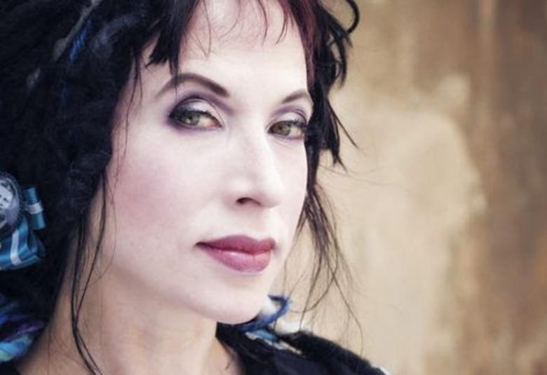 Kuvassa on Sofi Oksanen kasvokuvassa. Hänellä on tummat hiukset ja niissä vasemmalla pieni pyöreä kuva, jossa on miehen kasvoista kuva keskellä sini-musta-valkoista ruusuketta.