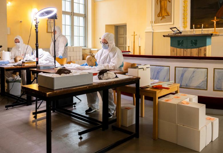 Kuvassa on etualalla tutkimuspöyt ja sen takana on henkilö valkoisessa suoja-asussa tekemässä tutkimusta. Taaempana vasemmalla on valkoisissa suoja-asuissa työskentelemässä pöydän ääeressä.