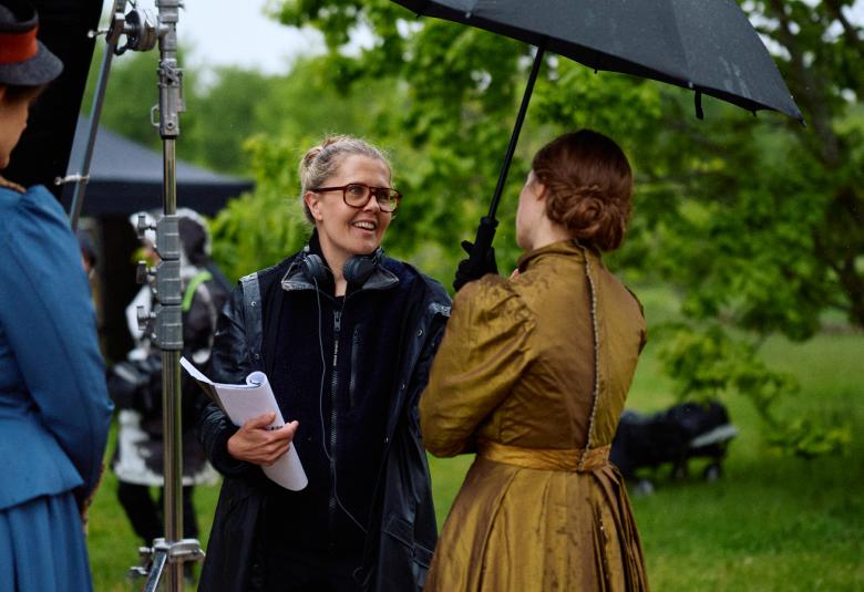 Kuvassa on Alli Haapasalo keskellä mustassa takissa selostamassa vihreässä vanhan aikaisessa puvussa olevalle näyttelijälle ja pitelee sateenvarjoa. Vasemmalla on sinisessä puvussa oleva näyttelijä katselee heitä. Tustalla on vihreitä puita.  