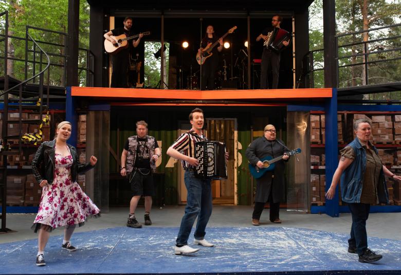 Kuvassa on näyttämö, jonka alaosassa on vasemmalla nuori nainen kesäisessä mekossa vauhdikkaassa asennossa ja hänen vieressään hanuristimies sekä oikealla nainen sinisessä farkkujakussa. Taustalla on kaksi miestä. Ylhäällä on parvi, jossa on kaksi kitaristia ja yksi hanuristi.