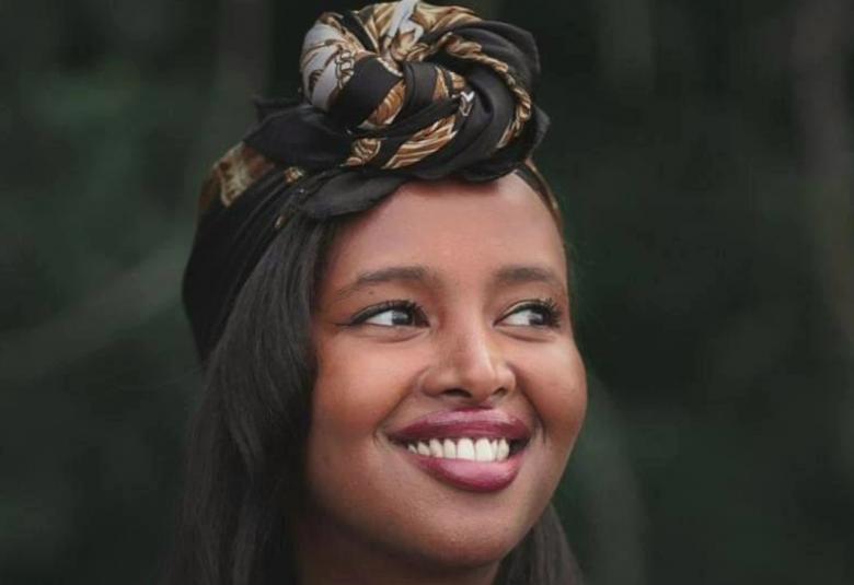 Kuvassa on Naima Mohamud on puolivartalokuvassa. Hänellä on tumma ihoja hänellä on musta ja vaaleakuvioinen huivi kiedottuna pään päälle. Hänellä on musta ja läpinäkyvähihainen pusero paällään. Tausta on tumma.