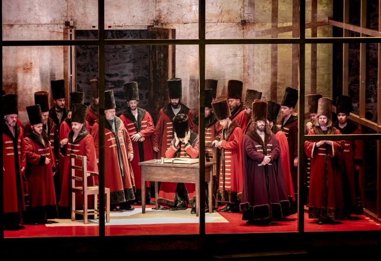 Kuvassa on punaisissa pitkissä takeissa ja joukko mustissa korkeissa hatuissa olevia henkilöitä seisomassa keskellä pöydän ääressä olevan miehen ympärillä.  Edessä on mustaa ikkunaristikkoa ja takana harmaata linnan muuria.