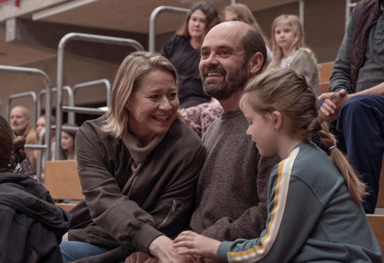 Kuvassa on vasemmalla vaaleahko nainen istumassa urheiluhallissa ja hänen vieressään istuu mies ja oikealla tyttö, jonka kanssa hän puhuu ja pitää kädestä kiinni.