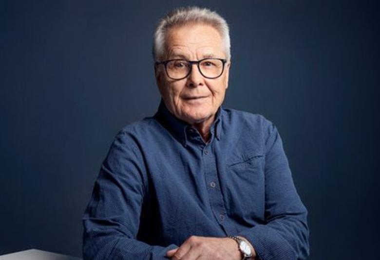 Kuvassa on Esa Siren istumassa kädet pöydällä ja hänellä on sininen paita päällään. Tausta on tumman sini-harmaa.