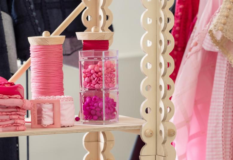 Kuvassa on vaalean punainen ja tumman punainen lankarulla, alas laskeutuva pitsinauha, taustalla on näkyvissä hiukan vaaleita ja vaalean punaisia vaatteita ripustettuna. 