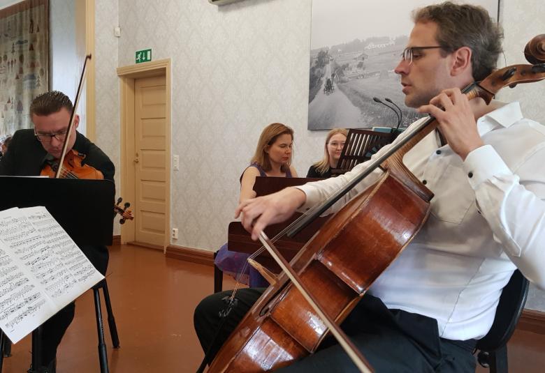 Kuvassa on taaempana soittamassa viulua Matti Koponen ja taaempana Liisa Malmivaara soittamassa flyygeliä.  Etualalla on Tuomas Ylinen valkoisessa paidassa soittamassa selloa nuotit edessään.