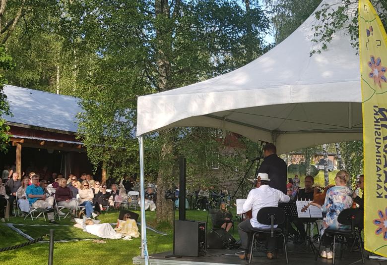 Kuvassa on etualalla valkoinen telttakatos, jonka alla on sottajia soittamassa ja laulaja selin. Oikeassa nurkassa keltainen leijamainos. Vasemmalla näkyy nurmikolla rinteessä yleisöä katsomassa konserttia. Takana on punainen katos, jossa on seinää auki. Taustalla on koivuja.