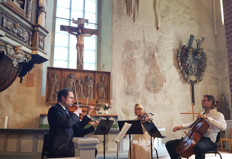 Kuvassa ovat vasemmalla Petteri Iivonen soittamassa viulua, takanaan Matti Koponen ja oikealla Eriikka Nylund vaaleassa mekossa soittamassa alttoviulua ja hänen oikealla puolella Tuomas Ylinen soittamassa selloa. Vasemmalla edessä näkyy osa saarnastuolista, joka on koristeltu kaiverruksin ja taustalla puinen alttarikaappi sekä sen yläpuolella krusifiksi. Seinällä on maalaukset ja hautausvaakuna.
