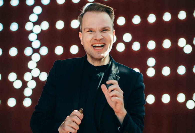 Kuvassa on nauravainen Kaapo Ijas seisomassa mustassa takissa ja paidassa. Taustalla valopalloja ruskeassa seinässä.