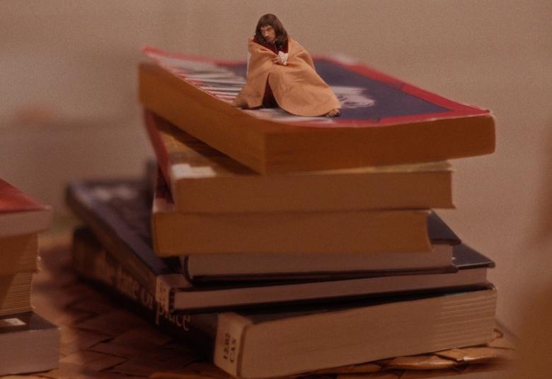 Kuvassa on viiden kirjan kirjapino ja sen päällä istuu vilttiin kääriytyneenä tummatukkainen nainen. Kuvan sävy on ruskehtava.