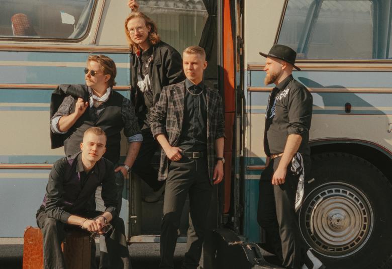 Kuvassa on Komiat yhtyeen miesjäsenet ryhmäkuvassa vaalean siniraitaisen bussin vieressä. Yksi istuu matkalaukun päällä ja yksi seisoo muita ylempänä keskellä.