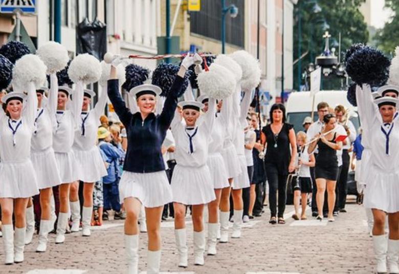 Kuvassa on kolmessa rivissä nuoria naisia marssimassa merimies-henkisissä, valkoisissa puolihamessa  ja puseroissa.  Heillä on kädet ylhääällä ja käsissä siniset tai valkoiset paperiset viuhkat.  Kadun molemmin puolin on yleisöä kadunvarsissa.