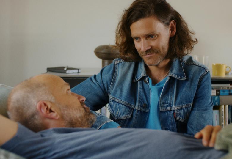 Kuvassa on sängyssä makaamassa sini-harmaassa paidassa ja hänen vieressään istumassa on mies sinisessä farkkutakissa. Hänellä on ruskeat pitkät hiukset.