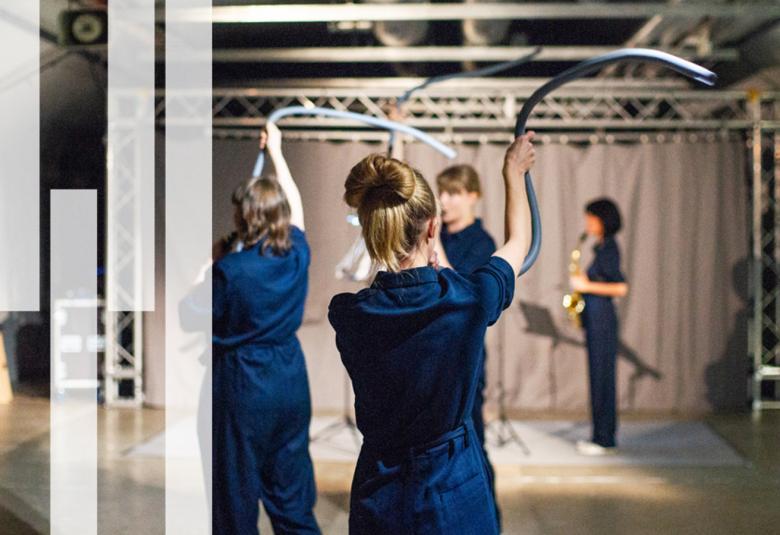 Kuvassa on tumman sinisissä housupuvuissa neljä nuorta naista, joista kolmella on kapeat muoviputket kädessään. Oikealla takana soittaa yksi nainen saksofonia.