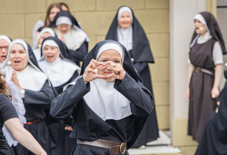 Kuvassa on keskellä nunna, joka näyttää käsillään sydäntä. Vasemmassa reunassa on nuori nainen, jolla ei ole nunnan asua. Takana molemmilla sivuilla on nunnia rivissä.