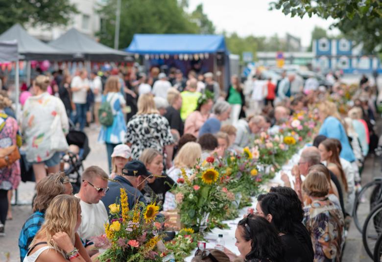 Kuvassa on yleisöä kesällä syömässä kesällä pitkän pöydän ääressä ja pöydällä on auringonkukkia. Taustalla kulkee yleisöä ja takana kaksi telttaa. 