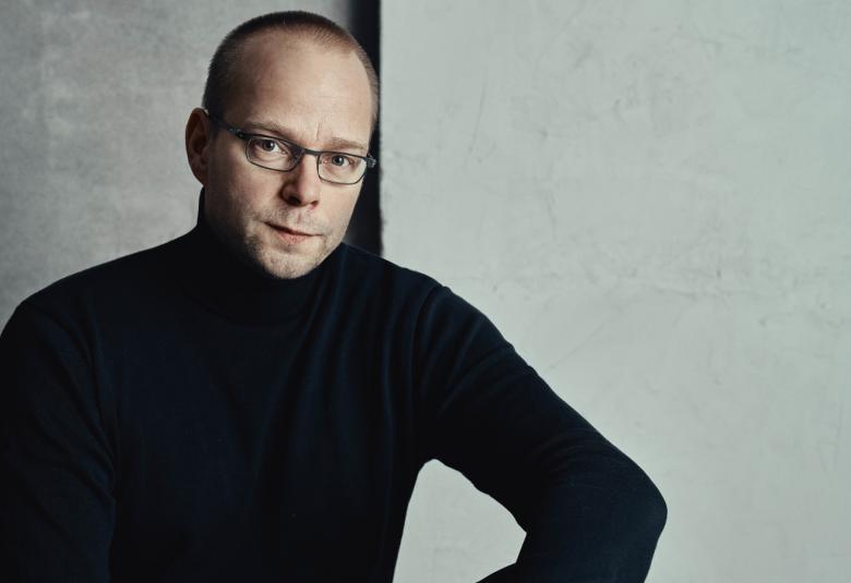 Kuvassa on Roope Lipasti istumassa ja hänellä on musta poolopaita päällään. Tausta on harmaa.