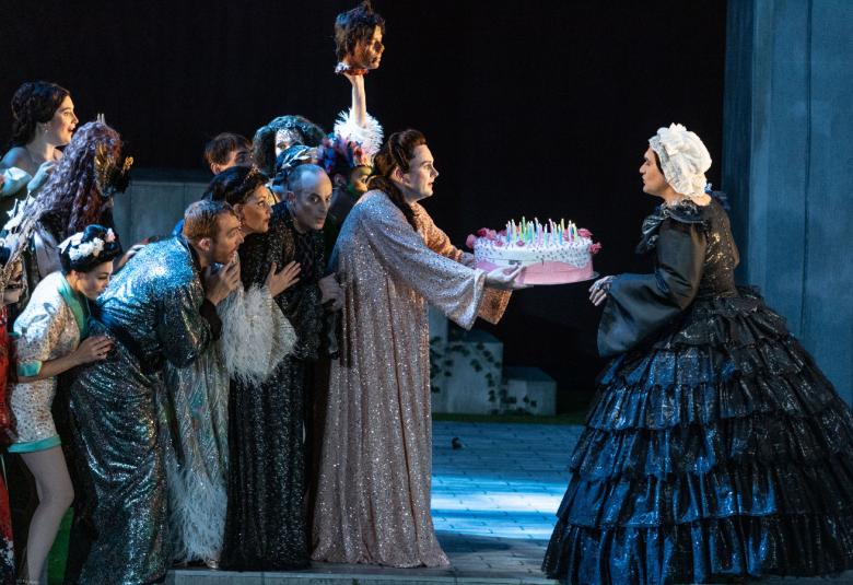 Kuvassa on vasemmalla joukko ihmisiä kasassa sivuttain katsomassa, kun etummainen mies ojentaa tumman sinisessä pönkkä hameessa olevalle naiselle täytekakkua, jossa on kynttilöitä, 