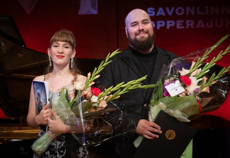  Kuvassa ovat puolivartalokuvassa vasemmalla Elsa Angervo isot kukat kädessään ja Martin Iivarinen myös samanlaiset kukat kädessään. Tausta on tumman punertava.