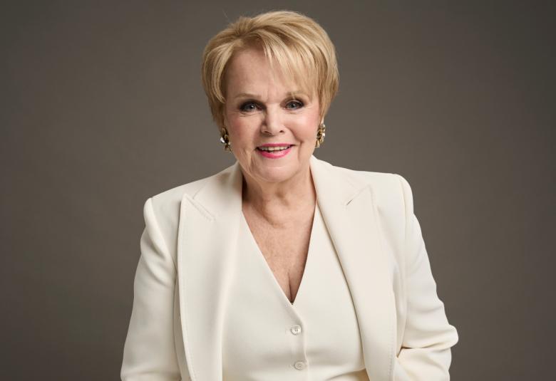 Kuvassa oN Katri Helenä valkoisessa takissa ja liivissä puolivartalokuvassa. Kuvan tausta on harmaa.