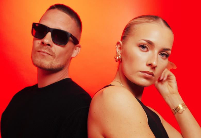 Kuvassa on puolivartalokuvassa vasemmalla miesaurinkolaseissa ja mustassa t-paidassa sekä nainen mustassa hihattomassa paidassa. Hänellä on vaaleat hiukset tiukasti päänmukaisesti. Tausta on voimakkaan oranssi.