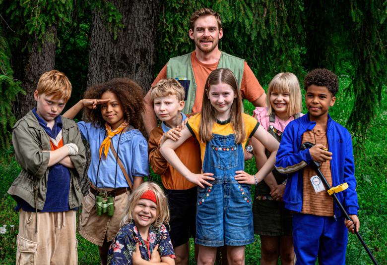 Kuvassa on seisomassa kesällä kuusi lasta ja istumassa yksi tyttö.  Takana seisoo Lauri Tilkanen. Taustalla on metsää. 