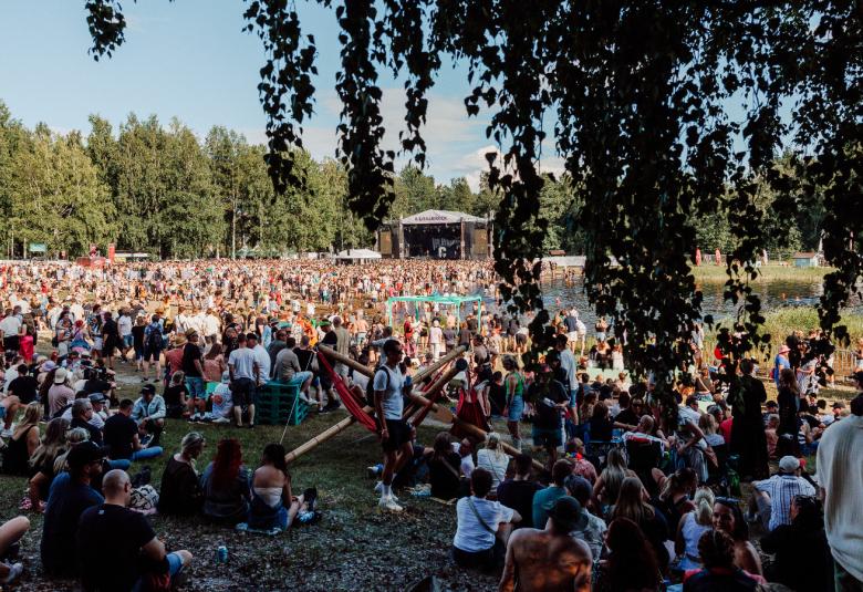 Kuvassa on kesäisessä säässä tuhansia ihmisiä tasaisella maalla ja taustalla on metsää. Edessä näkyy koivun oksia.