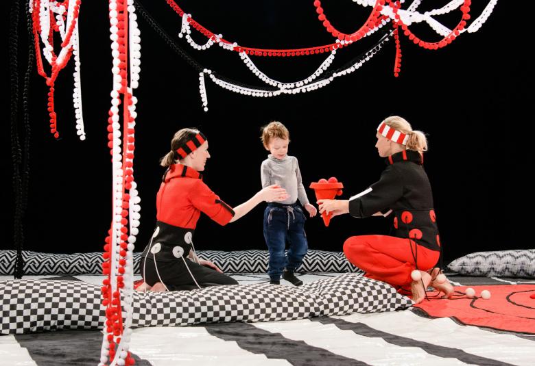 Kuvassa on näyttämöllä musta-valkoisia mattoja. Niiden päällä istuu kaksi naista ja heidän välissään pikkupoika. Yläpuolella ja sivussa roikkuu punaisia ja valkoisia nauhoja. Taustalla on mustaa.