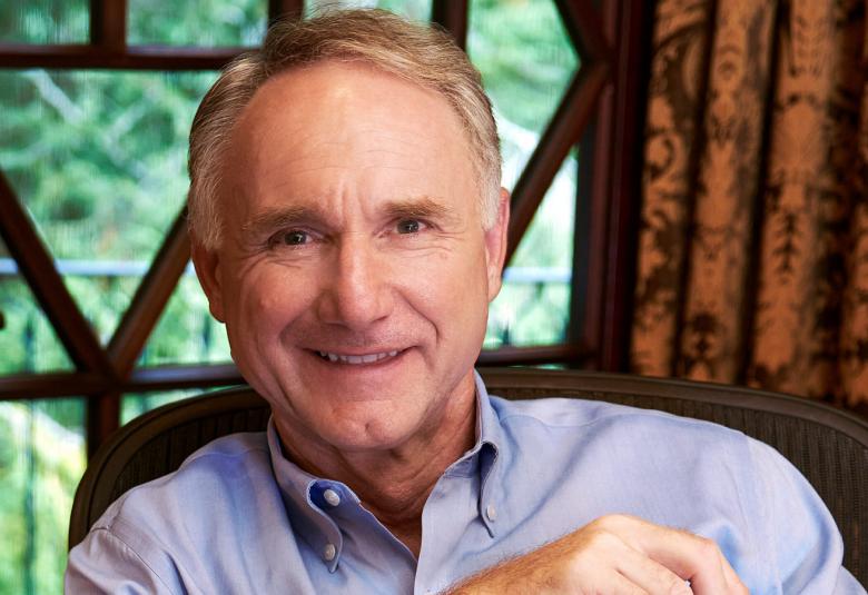 Kuvassa on Dan Brown puolivartalokuvassa. Hänellä on vaalean sininen kauluspaita päällään. Taustalla ikkunan takana vihreää ja oikealla ruskean mustaa verhoa. 