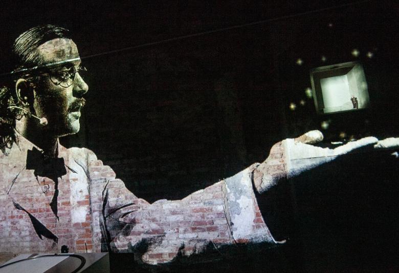 Kuvassa on Eetu Känkänen puolivartalokuvassa sivuttain pitämässä monitoria vasemmassa kädessään.  Hänellä on vaalean harmaa takki päällään ja tumma rusetti. Hänellä on metallipanta päässään.