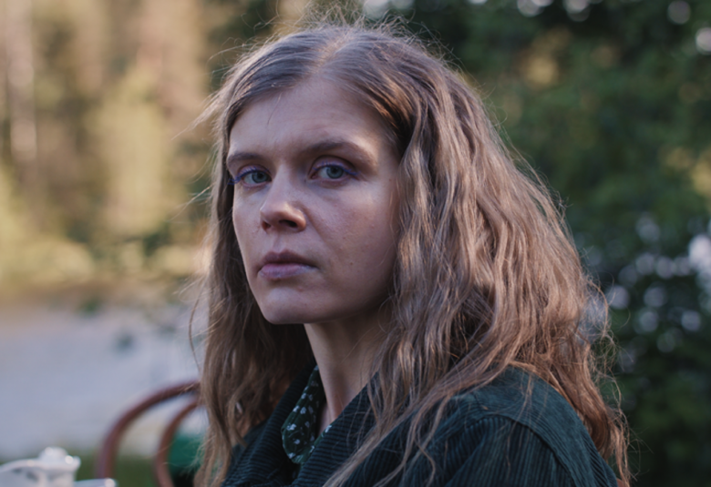 Kuvassa on Alina Tomnikov puolivartalokuvassa.  Hänellä on tumman sininen farkkutakki ja pitkät hiukset.  Taustalla on himmeästi järveä ja metsää.