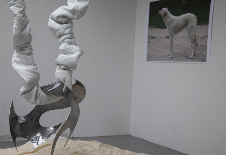 Kuvassa on vasemmalla teos, jossa on teräksinen kaarevista muodoista tehty jalusta ja sen päällä kaksi ylöspäin suuntautuvaa jalkaa ja niiden ympärillä olevaa vaaleata kangasta. Seinällä on suuren koiran kuva.