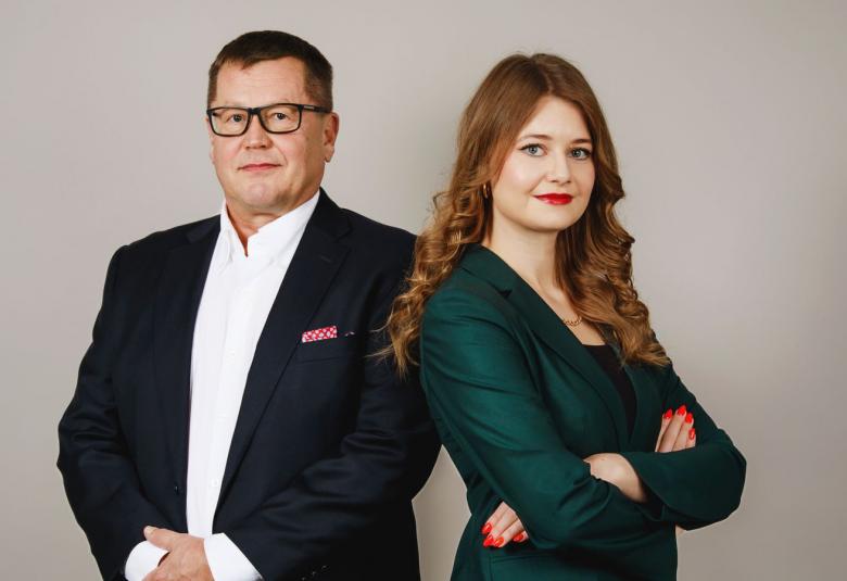 Kuvassa on vasemmalla Jukka Savolainen mustassa takissa ja valkoisessa paidassa. Oikealla on vinottain seisoo Iida Hallikainen ja hänellä on tumman vihreä jakku.  Kuvassa ovat he ovat puolivartalokuvassa. 