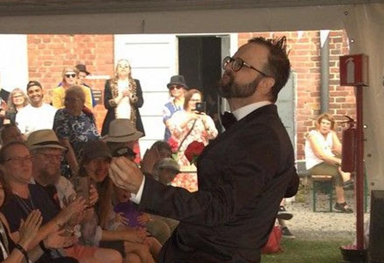 Kuvassa on oikealla Vellu Ristiniemi soittamassa sivuttain ilmakitaraa.  Edessä ja sivulla on yleisöä katsomassa esitystä seisoen ja istuen. 